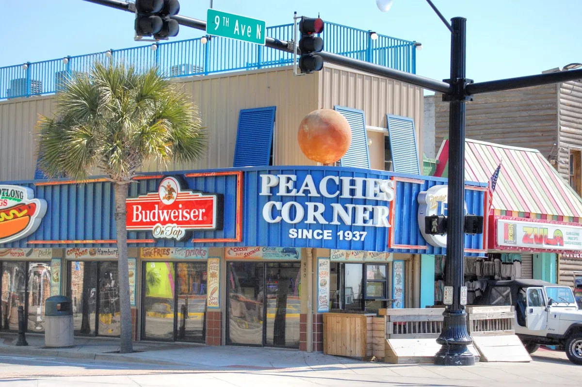 A vibrant street scene centered on the iconic Peaches Corner sign, with people dining and socializing beneath palm trees and festive decorations. The historic ambiance and energetic mood highlight Myrtle Beach’s local culinary and social experiences.