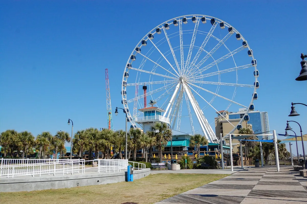 The iconic Myrtle Beach SkyWheel rises above palm trees and the lively boardwalk. Visitors stroll, dine outdoors, and savor the festive amusement park setting, with the Ferris wheel adding energy and excitement to the scene.
