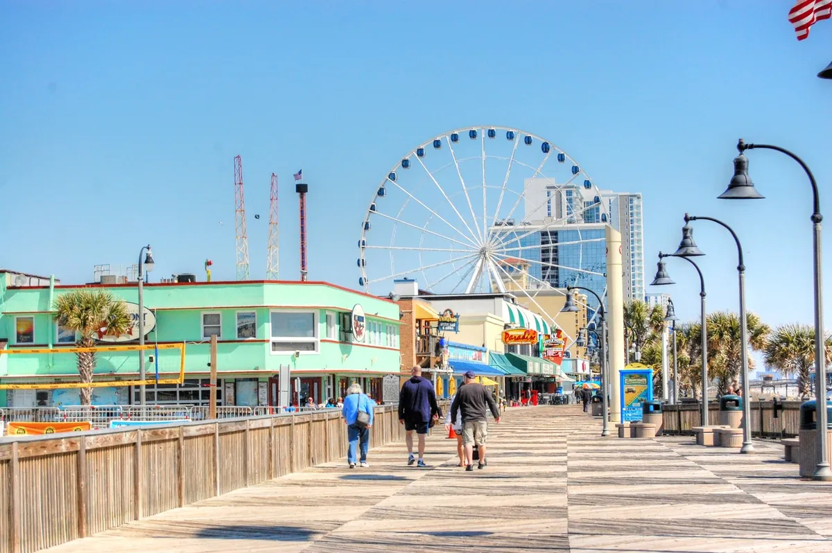 A lively seaside boardwalk scene featuring a Ferris wheel, colorful oceanfront buildings, and groups of people strolling along the wooden planks. The atmosphere is joyful and active, with opportunities for shopping, dining, and entertainment.
