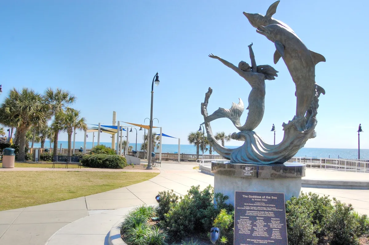 A whimsical beachfront park featuring the Goddess of the Sea sculpture, flanked by palm trees and park benches with the ocean as a backdrop. Visitors are seen strolling, taking photos, and enjoying the playful outdoor atmosphere.