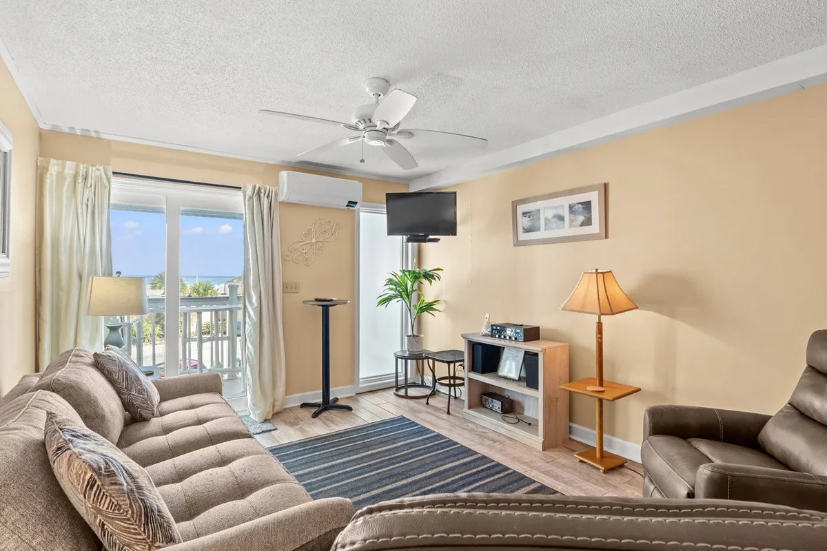 The image showcases a cozy living area with a comfortable sofa, a television for family entertainment, a warm lamp casting inviting light, and a vibrant potted plant adding a touch of freshness. The room feels open and welcoming, perfect for relaxing after a day on the beach, with hints of sunset light and a gentle, airy atmosphere.