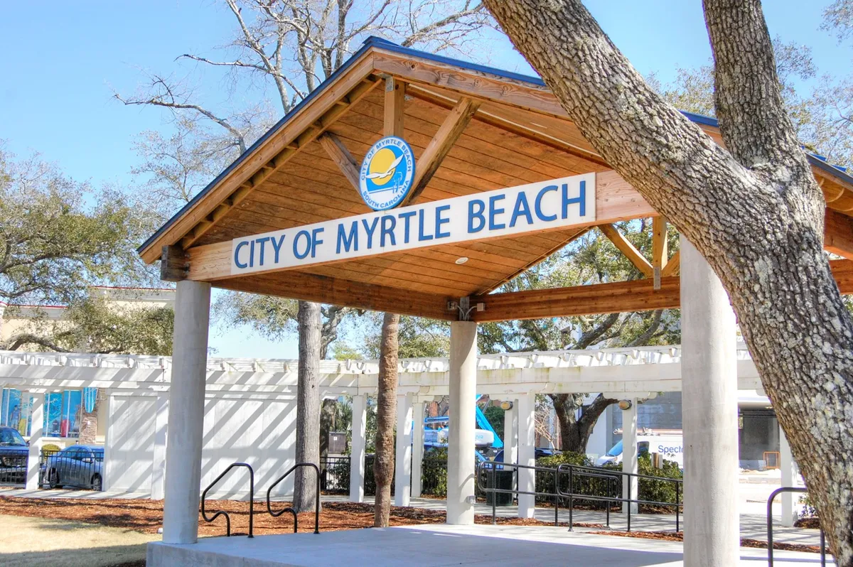 This image features the 'City of Myrtle Beach' sign beneath a wooden roof structure surrounded by trees, evoking the sense of arrival and access to local attractions and community events.