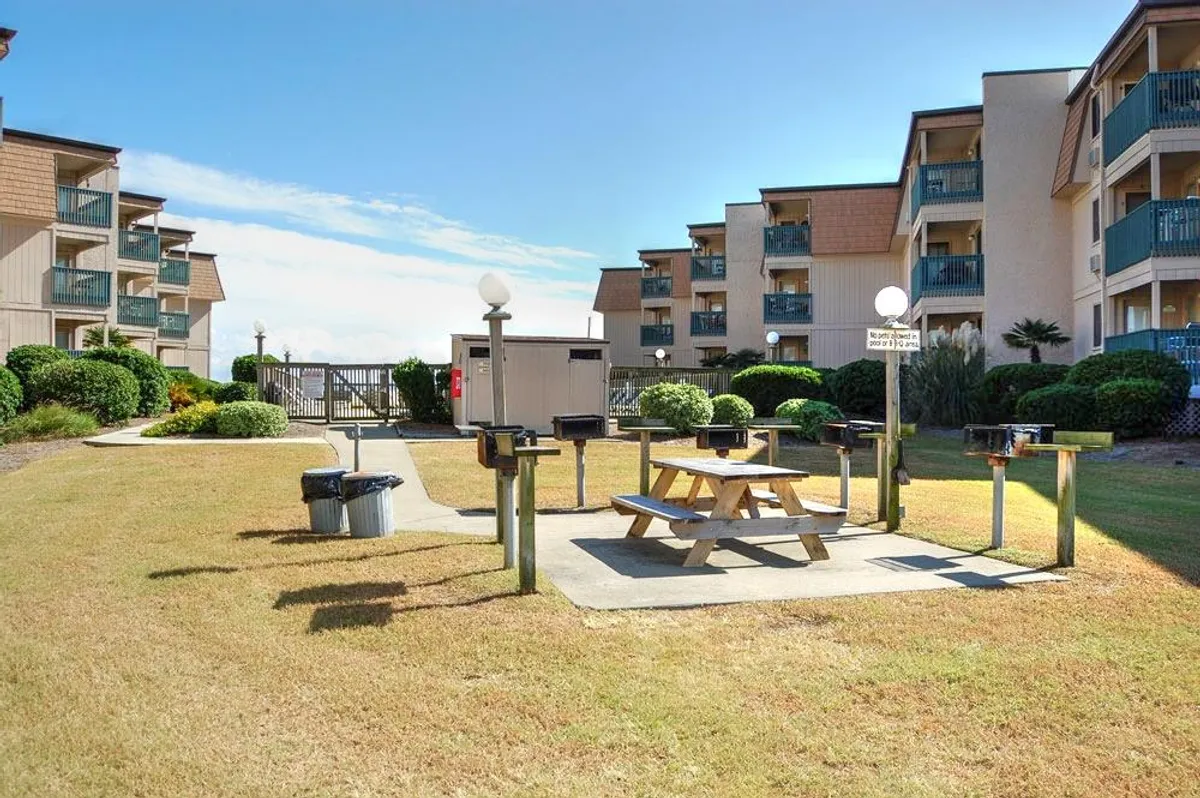 The photo shows a sunny grilling and picnic area featuring multiple grill stations, a picnic table, landscaped bushes, and the building's facade. The environment is casual and friendly, perfect for barbecues and social gatherings among guests.