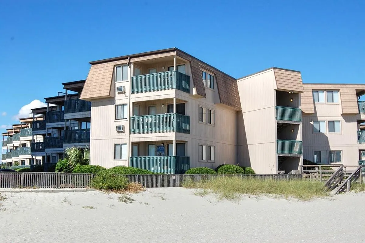 A welcoming beachfront condo building with private balconies overlooking the ocean, landscaped shrubs at the entrance, and a direct path leading to the sandy beach. The scene suggests ample space for gatherings and relaxation, with sunset glow illuminating the balconies.