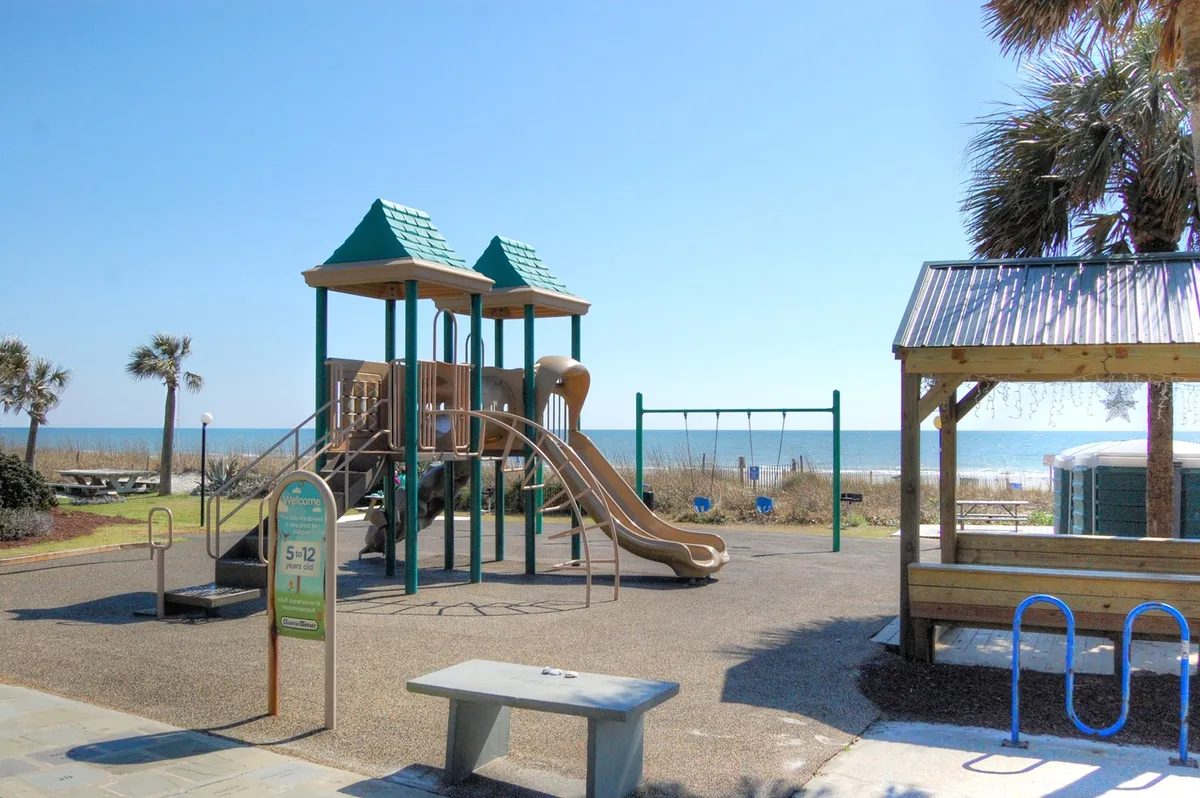 A vibrant playground with swings and play structures is set against a backdrop of the sparkling ocean. Families and children are seen enjoying the equipment, while adults relax in the shaded picnic area nearby. Palm trees and soft sand enhance the joyful, carefree beachside atmosphere.