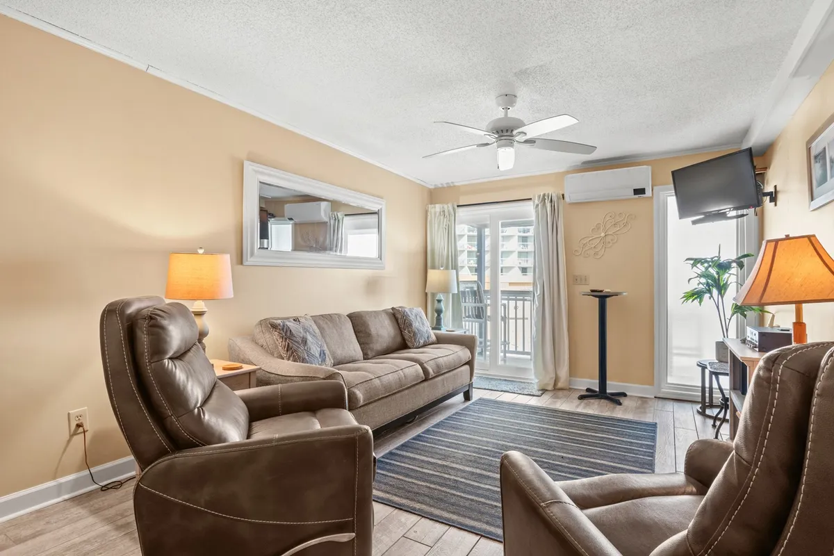 A cozy and inviting living room with a comfortable sofa, recliners, a lamp, mirror, curtains, and a television. Soft morning sunlight brightens the space, inviting guests to gather, socialize, watch TV, or relax together after a day at the beach.