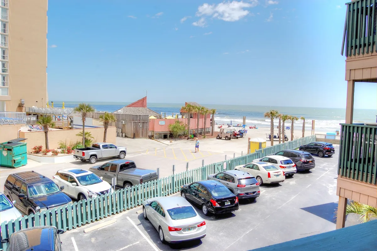 A scenic beach view with palm trees, a calm ocean, and sunlit sand, with a parking lot in the foreground and buildings in the distance. The setting is relaxed, ideal for beach outings, picnics, and sunset gatherings with the family.