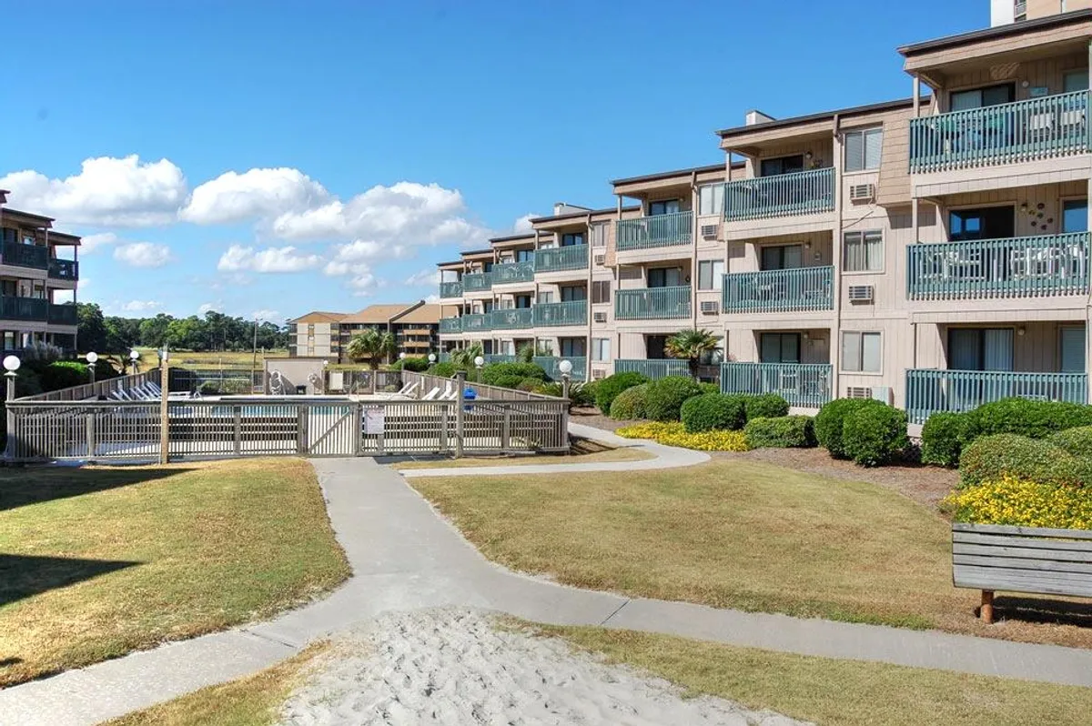 This exterior shot captures the resort grounds with landscaped gardens, a walkway leading to the pool area, and balconies of the apartments. The scene is relaxed and casual, suggesting easy access to amenities like the pool and barbecue area, and the atmosphere of casual leisure.